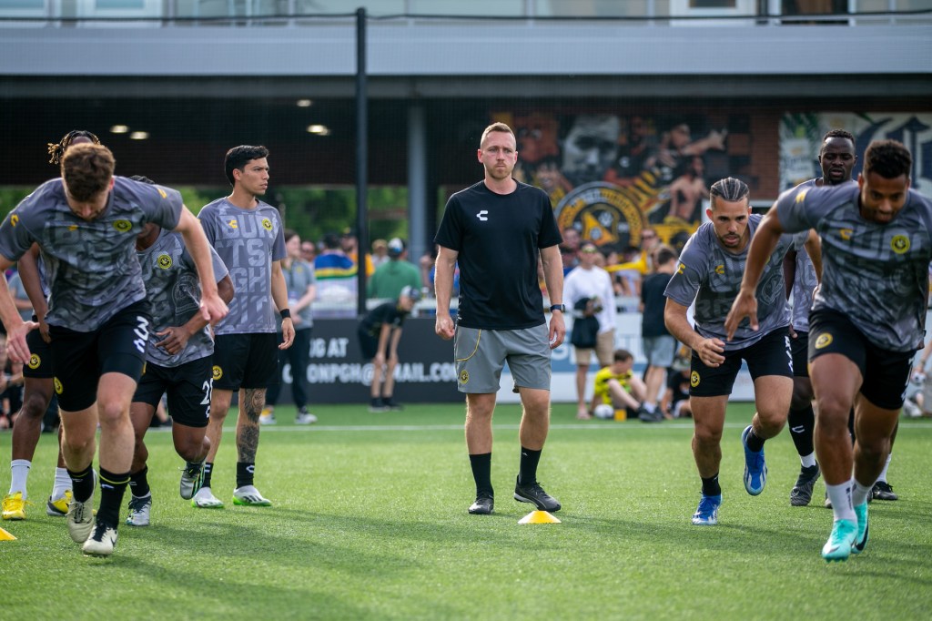 Hounds assistant coach Rob Vincent stands between lines of players during the team's warm-ups for the match against Indy Eleven on June 1, 2024 at Highmark Stadium. - Pittsburgh Riverhounds SC